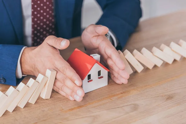 Cropped view of risk manager protecting house model from domino effect of falling wooden blocks with hands — Stock Photo