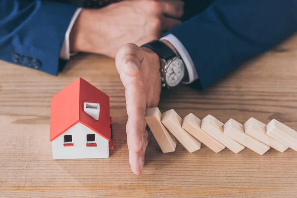 Cropped view of risk manager protecting house model from falling wooden blocks with hand — Stock Photo