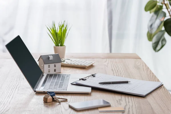 Scrivania in legno con computer portatile, smartphone, chiavi di casa, appunti con penna, modello di casa e pianta verde — Foto stock