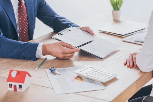 Cropped view of man giving clipboard and pen to businesswoman while sitting at desk — Stock Photo