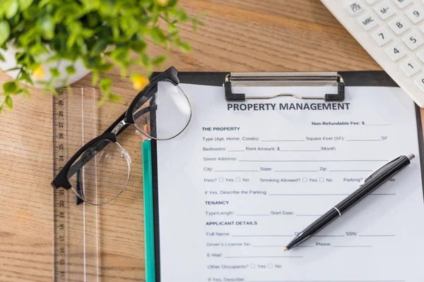 Top view of clipboard with property management agreement, pen, glasses, calculator and green plant on wooden desk — Stock Photo