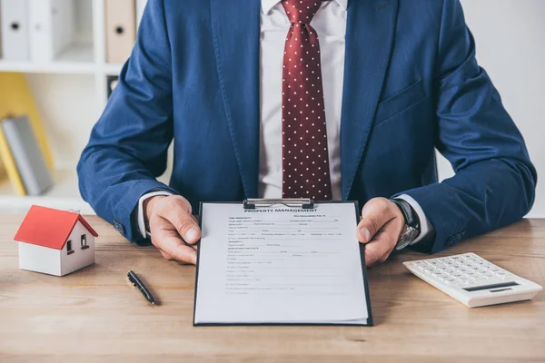 Vue partielle de l'homme d'affaires assis au bureau en bois près du presse-papiers, stylo, calculatrice et modèle de maison — Photo de stock