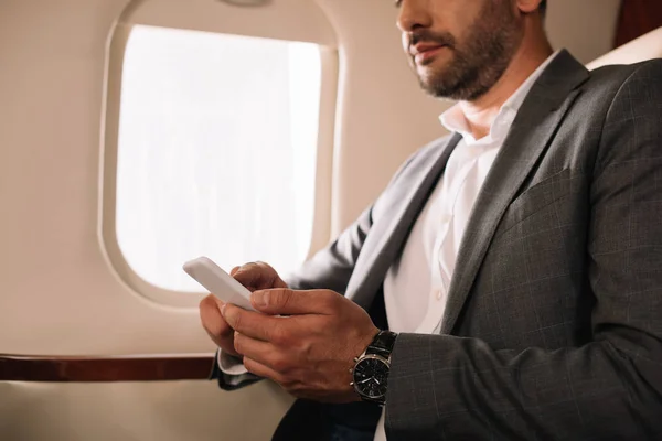 Vista recortada de hombre de negocios barbudo usando teléfono inteligente en jet privado - foto de stock