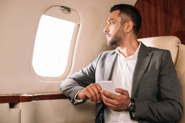 Hombre de negocios guapo mirando a la ventana del avión mientras se utiliza el teléfono inteligente en jet privado - foto de stock