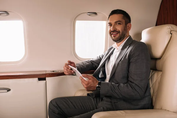 Cheerful bearded businessman in suit using smartphone in private jet — Stock Photo