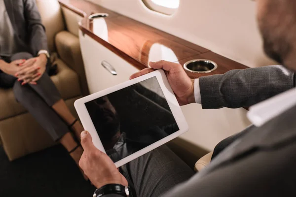 Vista recortada de empresario sosteniendo tableta digital con pantalla en blanco cerca de compañero de trabajo en jet privado - foto de stock