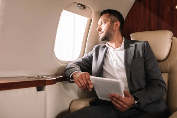 Bearded businessman in suit holding digital tablet while looking at airplane window in private jet — Stock Photo