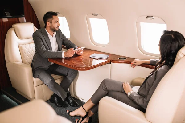 Selective focus of handsome bearded businessman holding gadget while sitting with businesswoman in private jet — Stock Photo