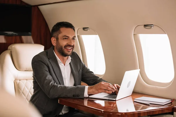 Enfoque selectivo de empresario barbudo feliz usando el ordenador portátil en primera clase - foto de stock