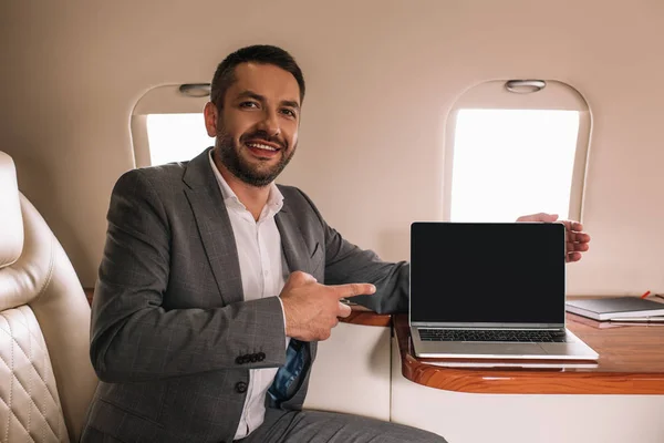 Hombre de negocios feliz señalando con el dedo a la computadora portátil con pantalla en blanco en jet privado - foto de stock