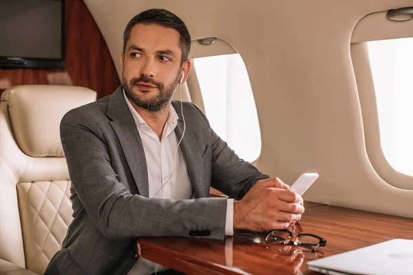 Selective focus of handsome businessman in earphones using smartphone in private jet — Stock Photo