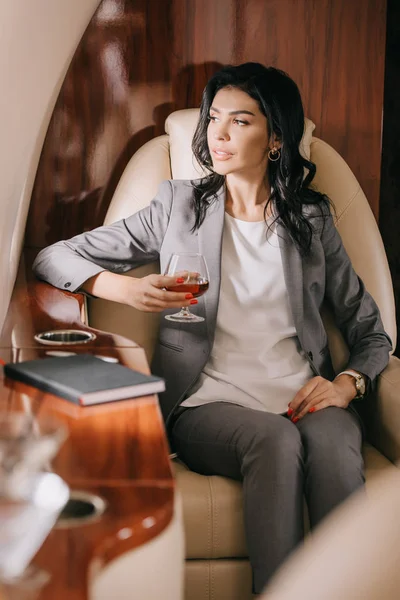Selective focus of businesswoman in formal wear holding glass with cognac in private jet — Stock Photo