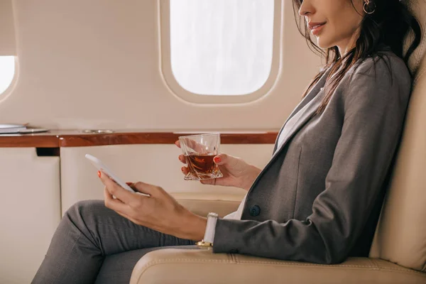 Vista recortada de la mujer de negocios en el desgaste formal celebración de vidrio con whisky y teléfono inteligente en jet privado - foto de stock