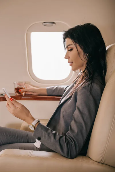 Біла жінка в офіційному одязі зі зі склом з віскі і смартфоном на приватному літаку. — стокове фото