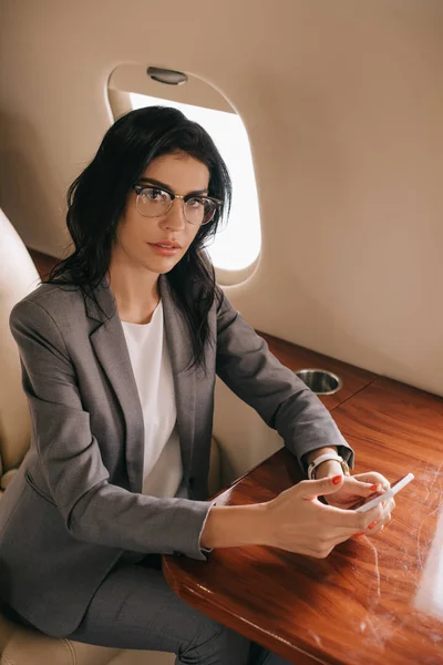 Vista aérea de atractiva mujer de negocios en gafas que sostienen el teléfono inteligente en jet privado - foto de stock