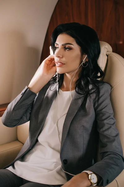 Attractive businesswoman touching earphones while listening music in private jet — Stock Photo