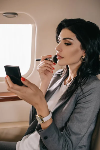 Attractive businesswoman holding cosmetic brush near face in private jet — Stock Photo