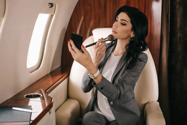 Messa a fuoco selettiva di attraente donna d'affari che tiene pennello cosmetico vicino al viso in jet privato — Foto stock