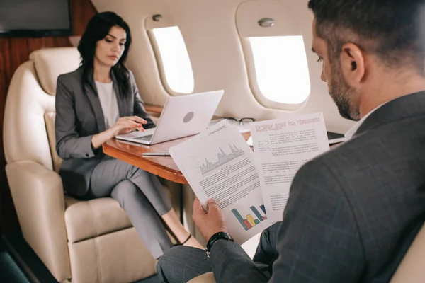Selektiver Fokus eines bärtigen Geschäftsmannes, der Diagramme und Grafiken in der Nähe einer attraktiven Geschäftsfrau mit Laptop im Flugzeug hält — Stockfoto