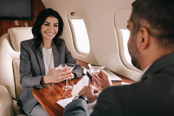 Селективний фокус щасливої бізнесменки і бородатого бізнесмена, який тримає окуляри Мартіні на приватному літаку. — стокове фото