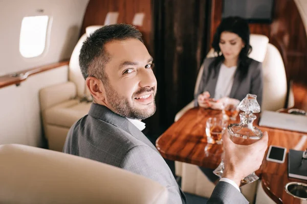 Selective focus of happy businessman holding jar with alcohol drink near businesswoman in private jet — Stock Photo