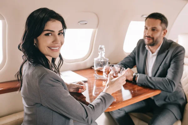 Enfoque selectivo de la mujer de negocios feliz sosteniendo vidrio con bebida alcohólica cerca de la mujer de negocios en jet privado - foto de stock