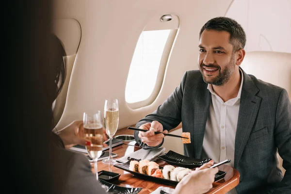 Vista recortada de la mujer de negocios cerca de alegre hombre de negocios y sabroso sushi en jet privado - foto de stock
