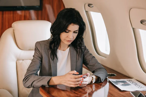 Attractive businesswoman holding smartphone near business newspaper in private jet — Stock Photo