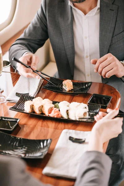 Обрезанный взгляд бизнесмена и бизнесвумен, держащих палочки для еды рядом с суши в частном самолете — стоковое фото