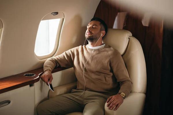 Handsome bearded man holding smartphone while listening music in private jet — Stock Photo