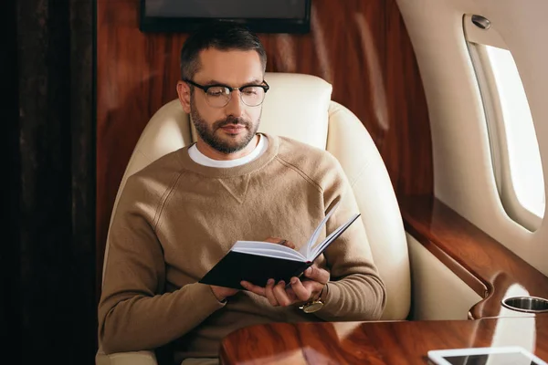 Bel homme barbu dans des lunettes regardant cahier en jet privé — Photo de stock