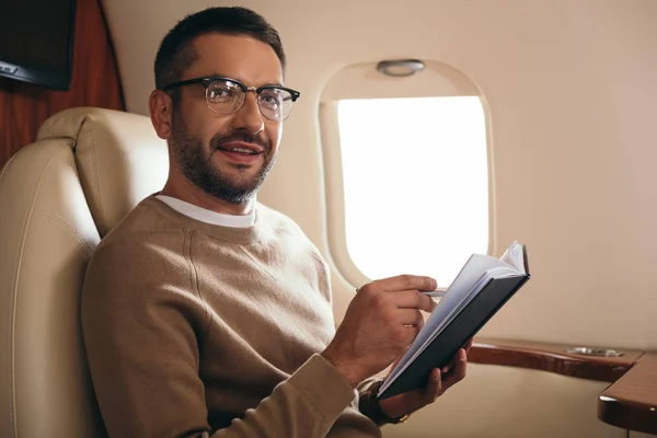 Hombre barbudo guapo en gafas mirando a la cámara mientras sostiene el cuaderno en jet privado - foto de stock