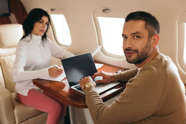 Enfoque selectivo de hombre feliz utilizando el ordenador portátil con pantalla en blanco cerca de la mujer en jet privado - foto de stock