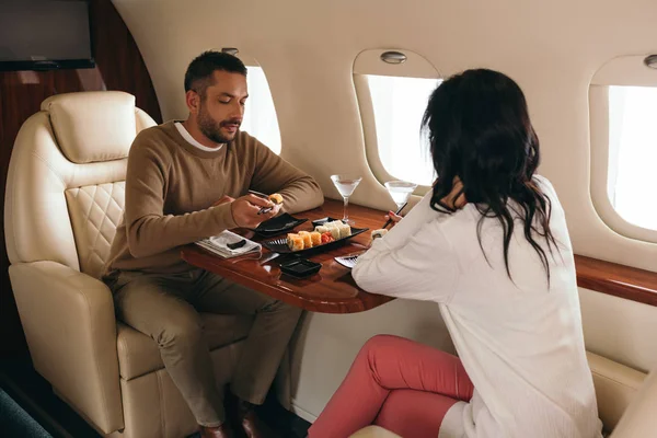 Вид сзади женщины, сидящей с мужчиной рядом с очками суши и мартини в частном самолете — стоковое фото