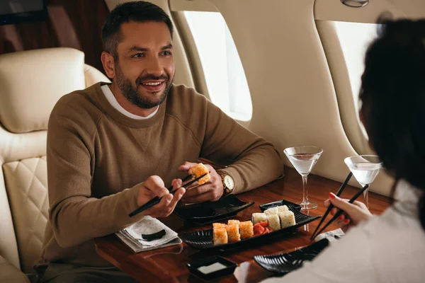 Обрезанный вид женщины, сидящей с счастливым мужчиной, держащим палочки для еды с рулоном в частном самолете — стоковое фото