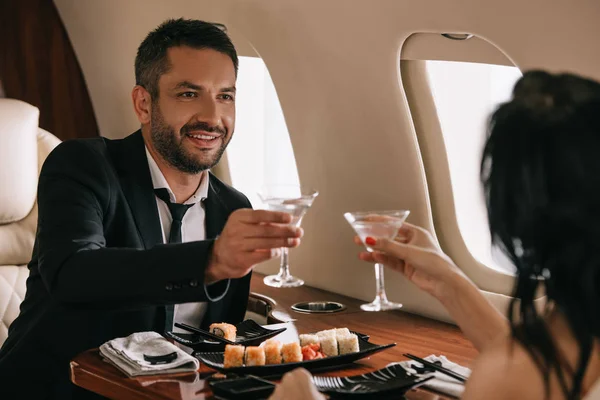 Vista recortada de la mujer sosteniendo vasos de martini cerca de hombre feliz en jet privado - foto de stock