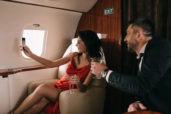 Happy man and woman in red dress holding martini glasses and taking selfie in private jet — Stock Photo