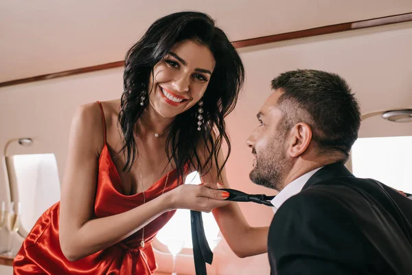 Selective focus of cheerful woman in red dress holding tie of bearded man in private jet — Stock Photo