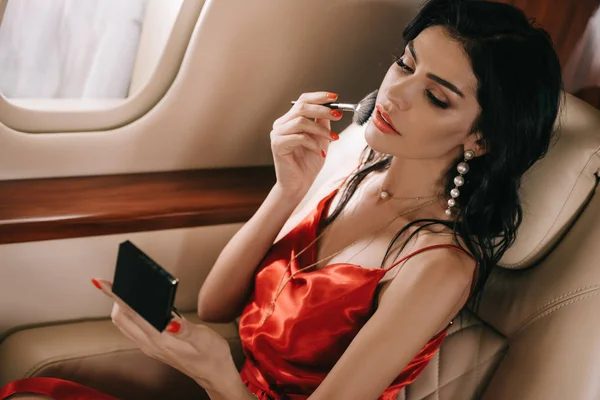 Привлекательная женщина в красном платье, нанося порошок для лица с косметической щеткой в частном самолете — стоковое фото