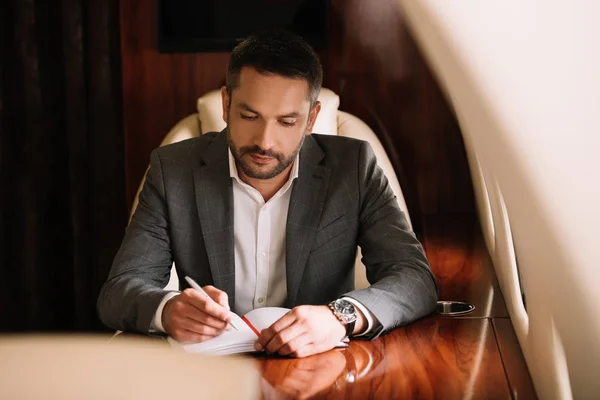 Selective focus of bearded businessman holding pen while writing in notebook and sitting in plane — Stock Photo