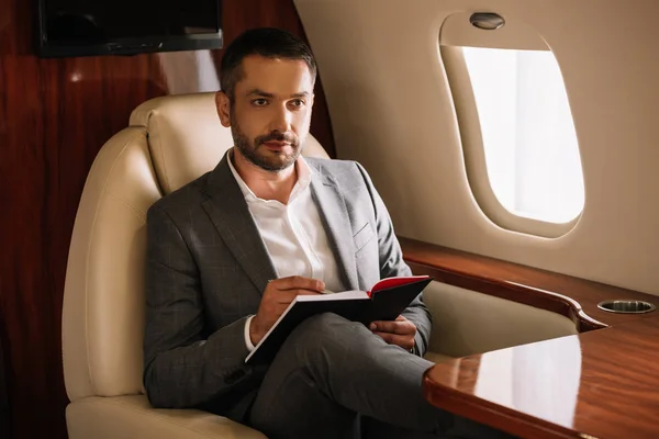 Selective focus of bearded businessman holding notebook and pen in private jet — Stock Photo