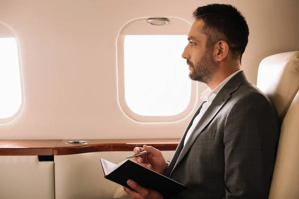 Profile of handsome bearded businessman holding notebook and pen in private jet — Stock Photo