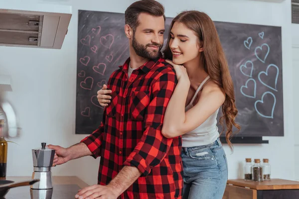 Attractive happy girl hugging smiling boyfriend making coffee in geyser coffee maker — Stock Photo