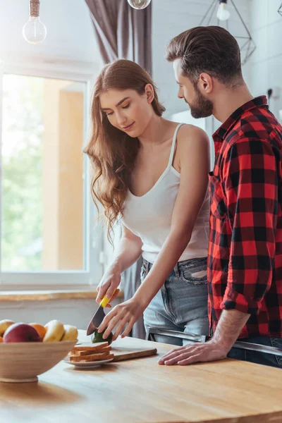 Junger Mann schaut schöne Freundin an, die frische Avocado zum Frühstück schneidet — Stockfoto