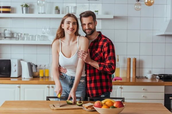 Feliz joven mujer cortando aguacate fresco y sonriendo a la cámara mientras sonríe novio abrazándola - foto de stock