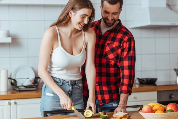 Улыбающийся мужчина, стоящий рядом с веселой девушкой, режет свежий авокадо — стоковое фото