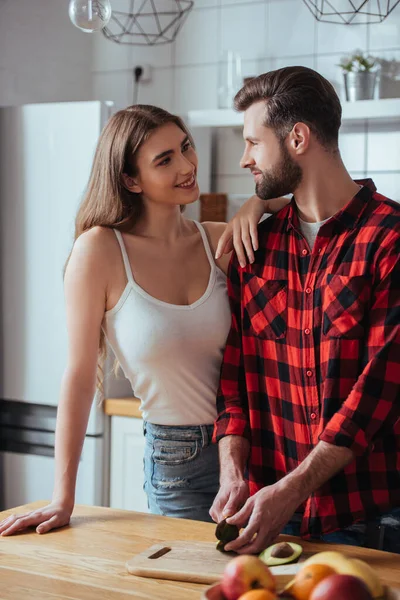 Feliz chica mirando guapo novio corte fresco aguacate cerca de cuenco con frutas frescas - foto de stock