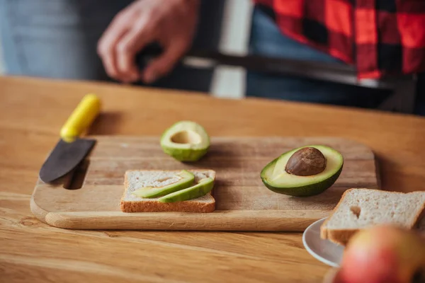 Cropped view of man near chopping board with bread, knife and halves of fresh avocado — Stock Photo