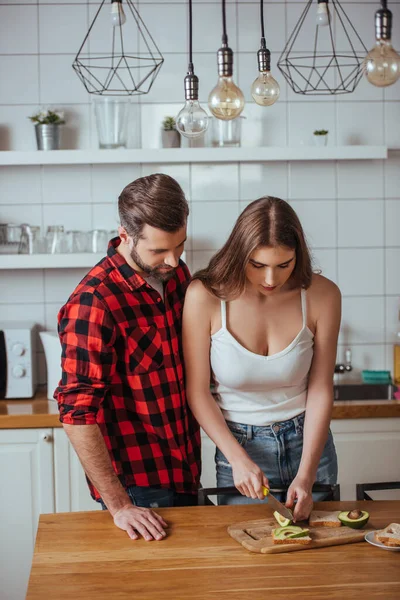 Jovem olhando para namorada atraente cortando abacate fresco para o café da manhã — Fotografia de Stock