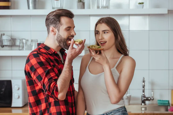 Молода пара тримає тости зі свіжим авокадо, стоячи на сучасній кухні — стокове фото